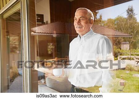 Stock Image - Senior man at home, drinking coffee, looking out of window. Fotosearch