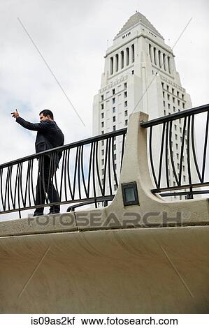 Stock Image - Side view of business man standing on walkway, hand raised, Los Angeles City Hall, California, USA. Fotosearch