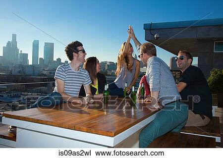 Stock Image - Six adult friends drinking beer at table of rooftop bar with Los Angeles skyline, USA. Fotosearch