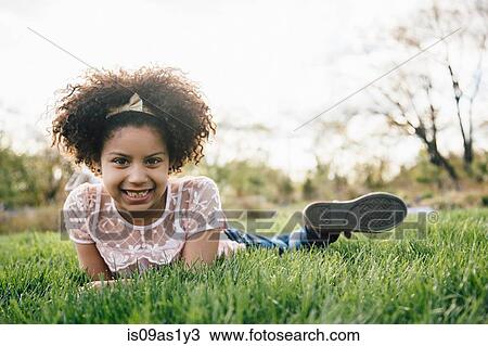 Stock Image - Surface level view of girl lying on front on grass smiling, looking at camera. Fotosearch