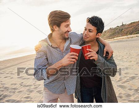 Stock Image - Two male friends having drinks together on beach, arms around each other. Fotosearch