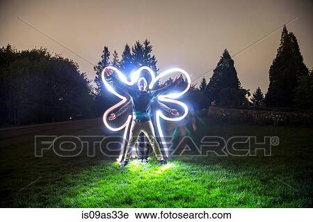 Stock Image - Two men standing together in field at dusk, creating star shape with bodies, friend tracing body shape with light trail. Fotosearch