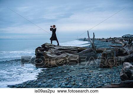 Stock Photograph - Woman looking through binoculars from large driftwood tree stump on beach. Fotosearch
