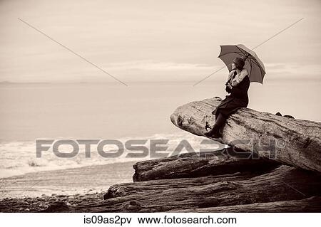 Stock Photo - Woman with umbrella sitting on large driftwood tree trunk on beach. Fotosearch