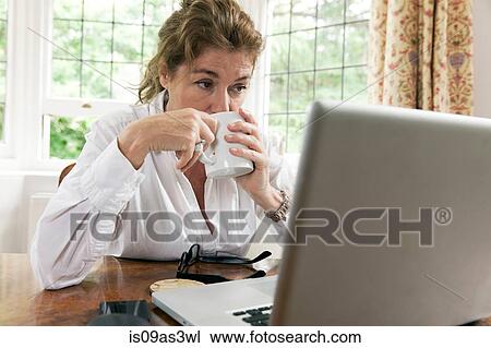 Stock Image - Worried mature woman working on laptop whilst drinking coffee in living room. Fotosearch