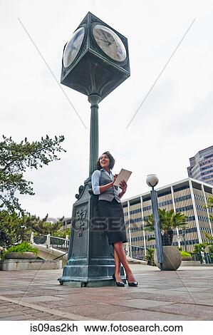 Stock Image - Young business woman leaning against clock using digital tablet, looking away. Fotosearch