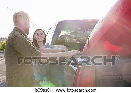 Stock Photo - Young couple leaning against pick up truck, looking at each other smiling, lens flare. Fotosearch