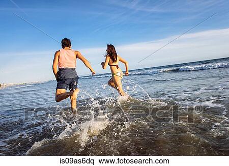 Stock Photography - Young couple running in sea, rear view. Fotosearch