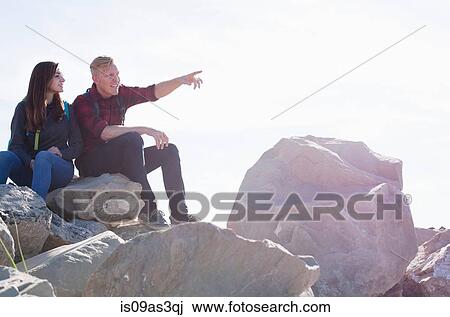 Stock Image - Young couple sitting on rocks looking away pointing. Fotosearch
