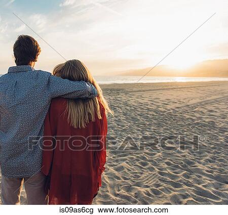 Stock Image - Young couple standing on beach, hugging, rear view. Fotosearch