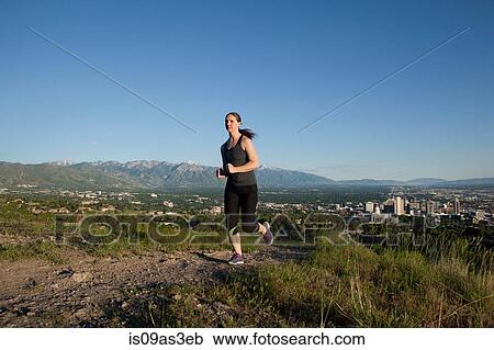 Stock Image - Young female runner running along track above city in valley. Fotosearch