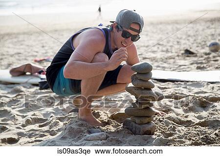 Young man crouching next to a stacked stones on beach View Large Photo Image Stock Image - Young man crouching next to a stacked stones on beach. Fotosearch