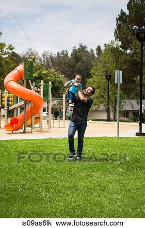 Stock Image - Young man playing holding up toddler brother in park. Fotosearch