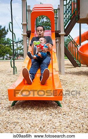 Stock Image - Young man sliding down with toddler brother on playground slide. Fotosearch