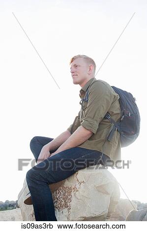 Stock Image - Young man wearing backpack sitting on rock looking away. Fotosearch