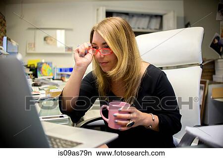 Young woman in office, sitting at desk, holding coffee cup, putting on glasses View Large Photo Image Picture - Young woman in office, sitting at desk, holding coffee cup, putting on glasses. Fotosearch