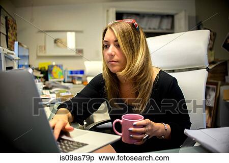 Young woman in office, sitting at desk, holding coffee cup, using laptop View Large Photo Image Stock Photography - Young woman in office, sitting at desk, holding coffee cup, using laptop. Fotosearch