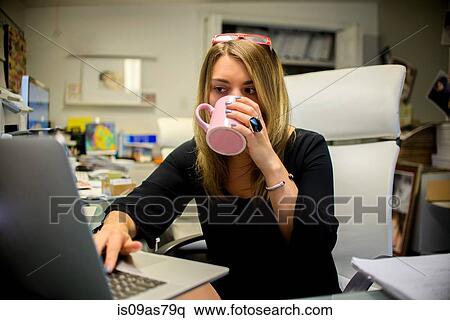 Stock Photography - Young woman in office, sitting at desk, drinking coffee, using laptop. Fotosearch