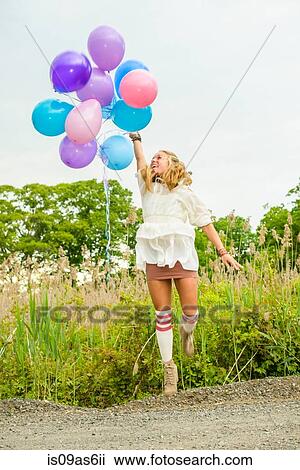 Stock Image - Young woman jumping with bunch of balloons on rural road. Fotosearch