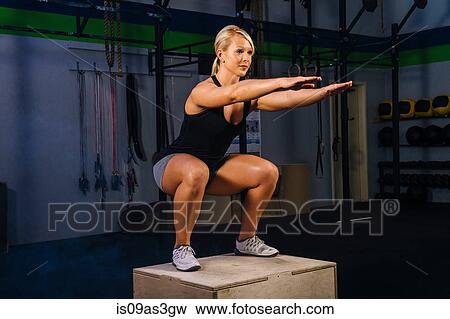 Young woman squatting on gym box with arms reaching out View Large Photo Image Stock Photo - Young woman squatting on gym box with arms reaching out. Fotosearch