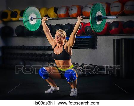 Stock Image - Young woman squatting with barbell raised in gym. Fotosearch
