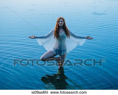 Stock Image - Young woman standing in blue lake practicing yoga. Fotosearch