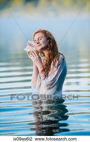 Stock Image - Young woman standing in misty lake with eyes closed listening to seashell. Fotosearch