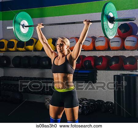 Stock Image - Young woman weight lifting with barbell in gym. Fotosearch