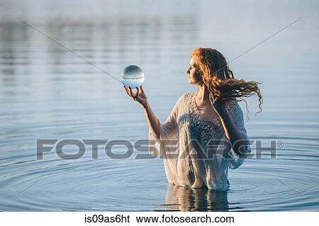 Stock Photograph - Young woman with long red hair standing in lake gazing at crystal ball. Fotosearch