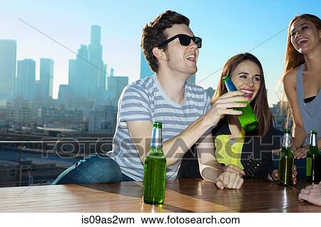 Picture - Young women and man drinking beer and laughing at rooftop bar with Los Angeles skyline, USA. Fotosearch