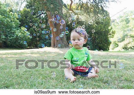 Picture - Baby boy sitting on grass watching bubbles in park. Fotosearch