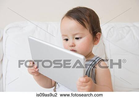 Stock Image - Baby boy sitting on sofa holding up digital tablet. Fotosearch