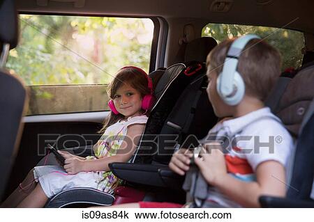 Stock Image - Boy and younger sister wearing headphones and using digital tablet in car back seat. Fotosearch