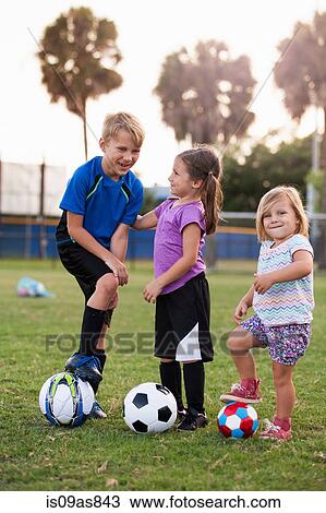 Stock Image - Boy and younger sisters with footballs on practice pitch. Fotosearch