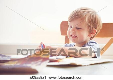 Stock Image - Boy sitting at table holding crayons, looking away smiling. Fotosearch