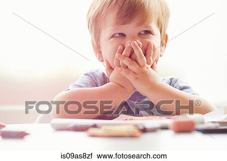 Stock Image - Boy sitting at table resting on elbows, covering mouth with hands, looking away. Fotosearch