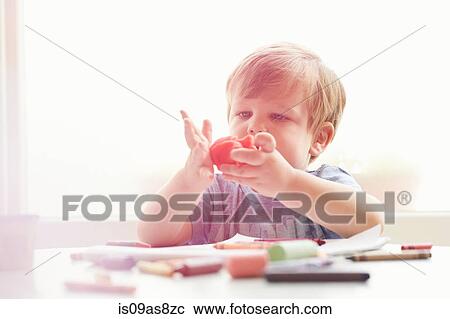 Stock Image - Boy sitting at table with art supplies playing with modelling clay. Fotosearch