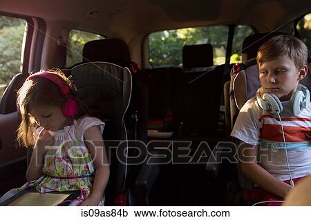Stock Image - Boy watching younger sister using digital tablet in car back seat. Fotosearch
