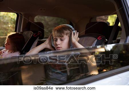 Stock Image - Boy with younger sister listening to headphones in car back seat. Fotosearch