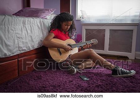 Stock Image - Girl sitting on bedroom floor playing guitar looking down. Fotosearch