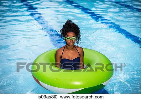 Stock Image - Girl standing in inflatable ring in swimming pool, wearing goggles looking at camera. Fotosearch