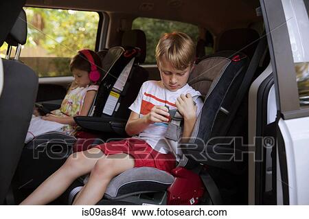 Stock Image - Girl using digital tablet whilst brother fastens seat belt in car back seat. Fotosearch