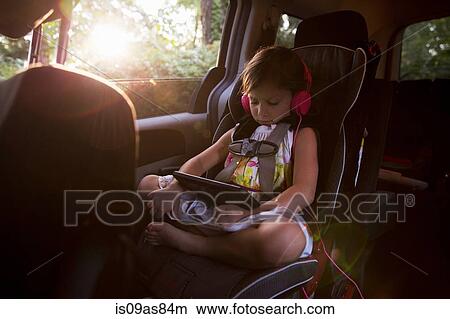 Picture - Girl wearing headphones using digital tablet in car back seat. Fotosearch