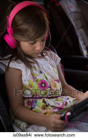 Stock Image - Girl wearing pink headphones concentrating on digital tablet in car. Fotosearch