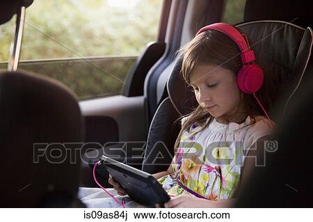 Stock Image - Girl wearing pink headphones using digital tablet in car. Fotosearch