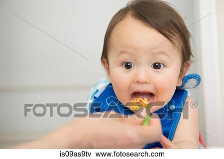 Hand of mother feeding baby food to open mouthed son in kitchen View Large Photo Image Stock Photo - Hand of mother feeding baby food to open mouthed son in kitchen. Fotosearch