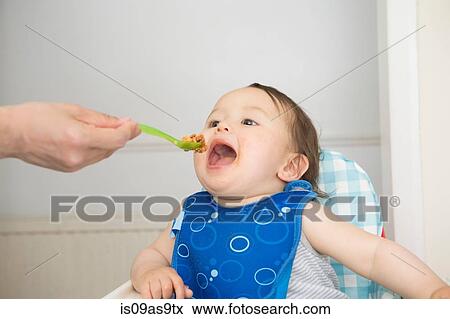 Hand of mother feeding baby food to open mouthed son in kitchen high chair View Large Photo Image Stock Photo - Hand of mother feeding baby food to open mouthed son in kitchen high chair. Fotosearch