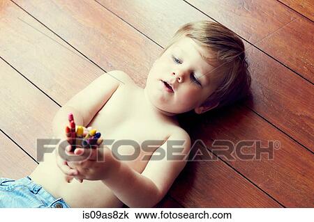 Stock Image - High angle view of boy lying on back on wooden floor holding crayons. Fotosearch