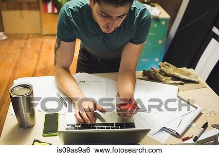 Stock Photograph - High angle view of young man using credit card and laptop to make online purchase. Fotosearch