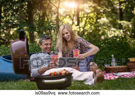 Stock Photography - Mature man and girlfriend looking at digital tablet whilst having picnic barbecue. Fotosearch